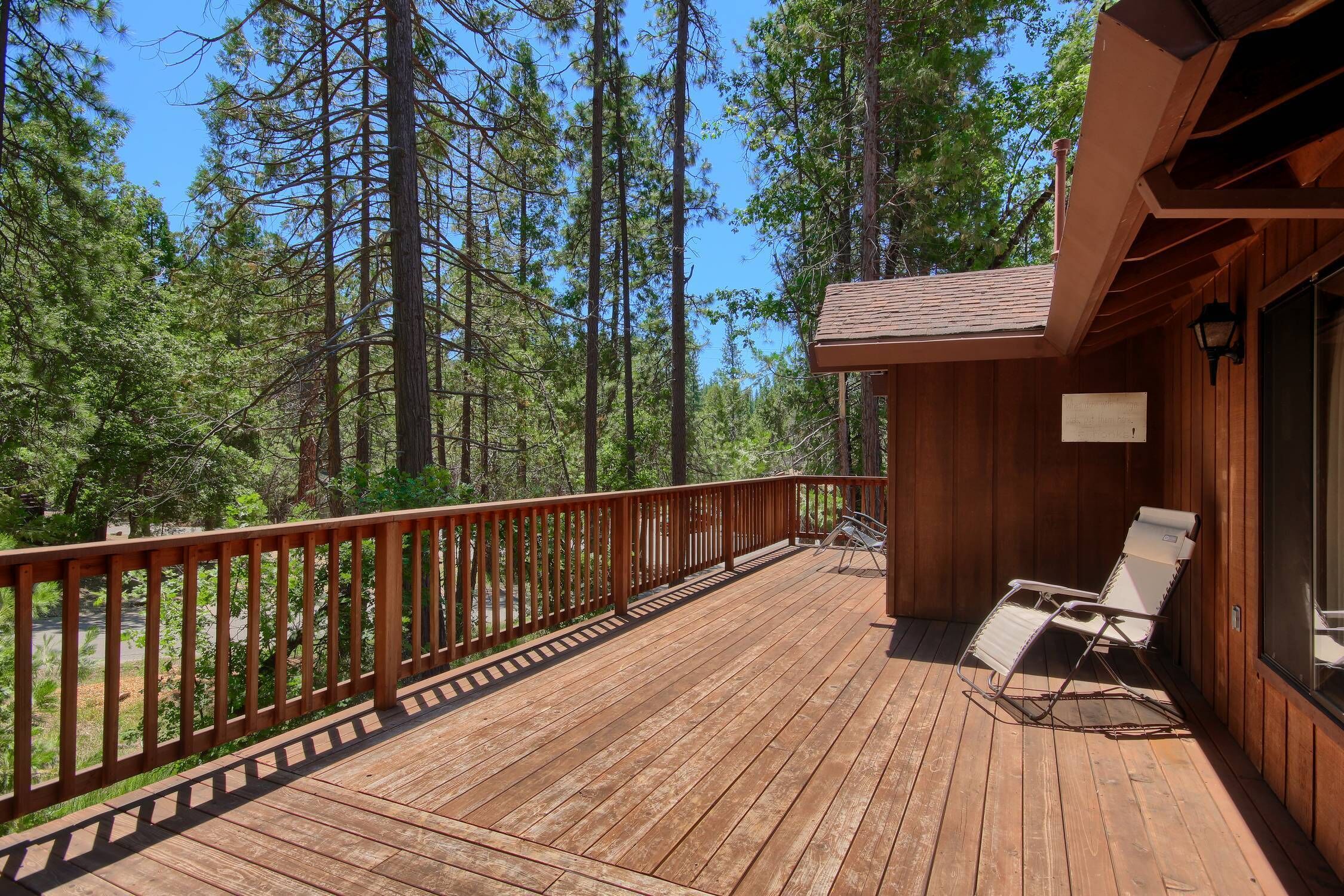 A wooden deck with a railing, two lounge chairs, and a view of a forest with tall trees.