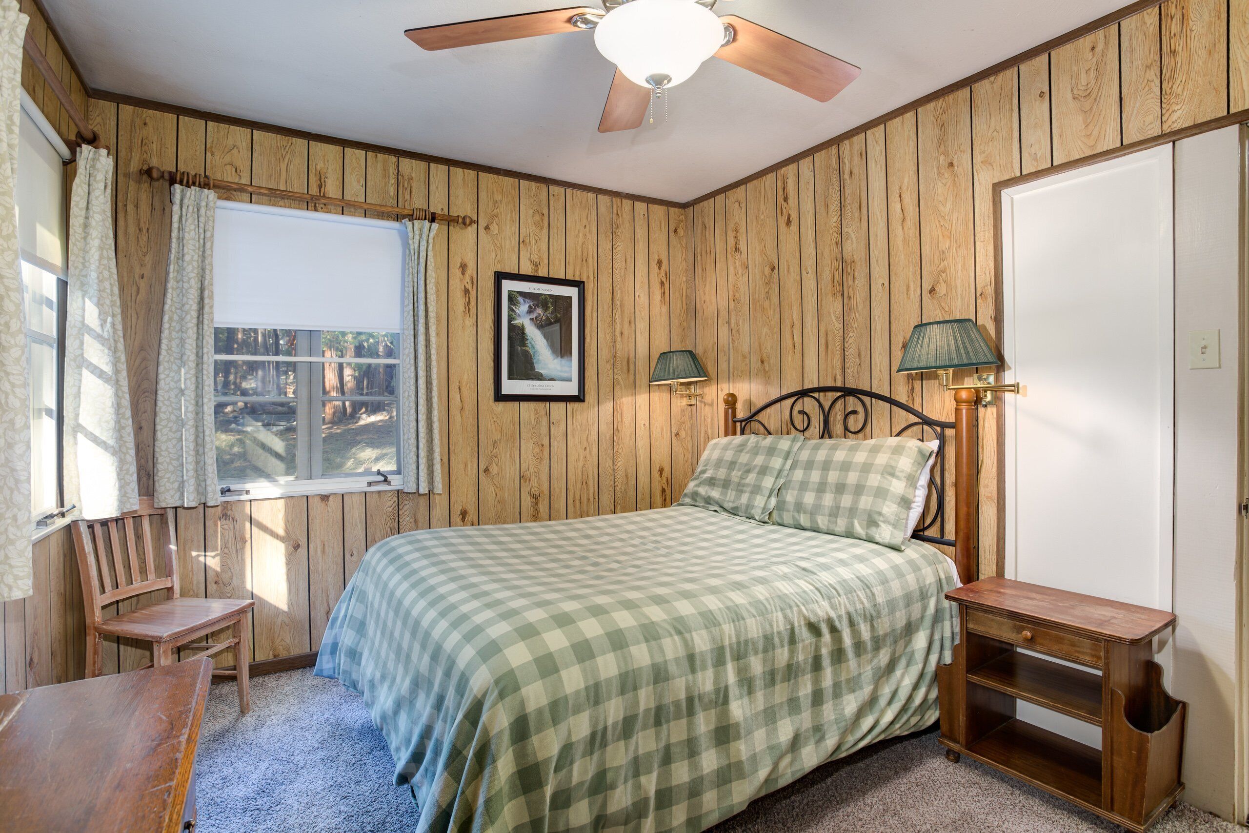 A bedroom with wooden panel walls, a bed with a checkered bedspread, a nightstand with a lamp, a chair, and a ceiling fan.