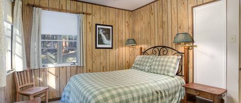 A bedroom with wooden panel walls, a bed with a checkered bedspread, a nightstand with a lamp, a chair, and a ceiling fan.