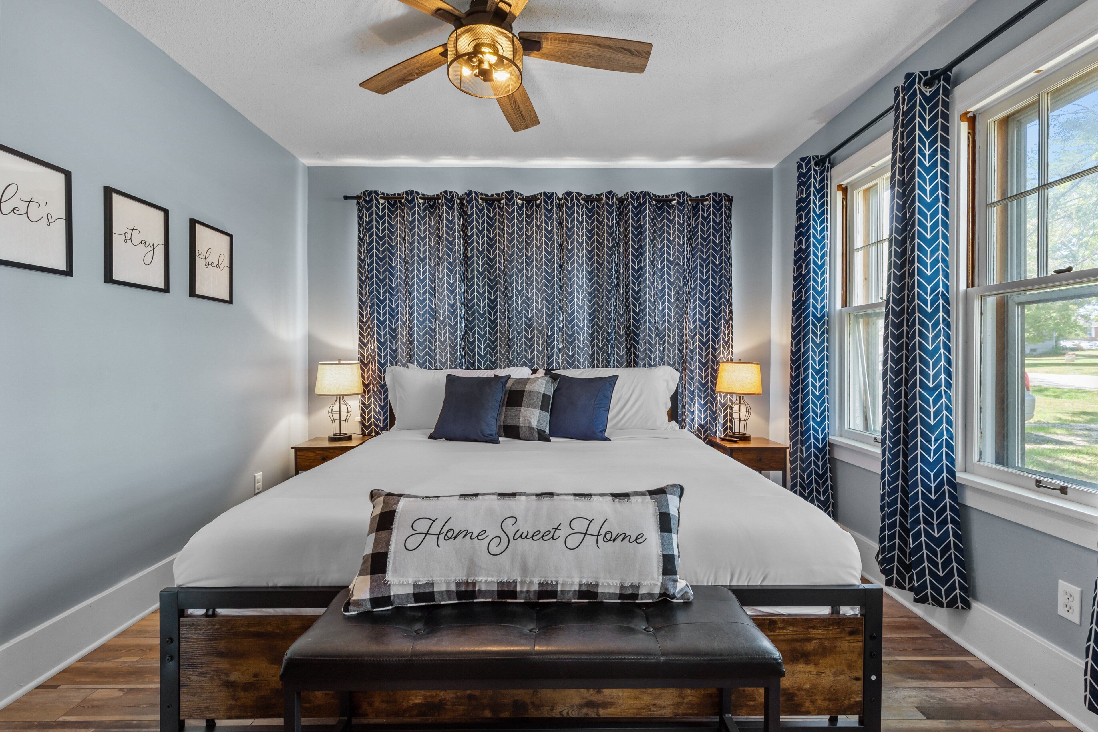 A bedroom with a bed, a bench, a ceiling fan, and patterned curtains.