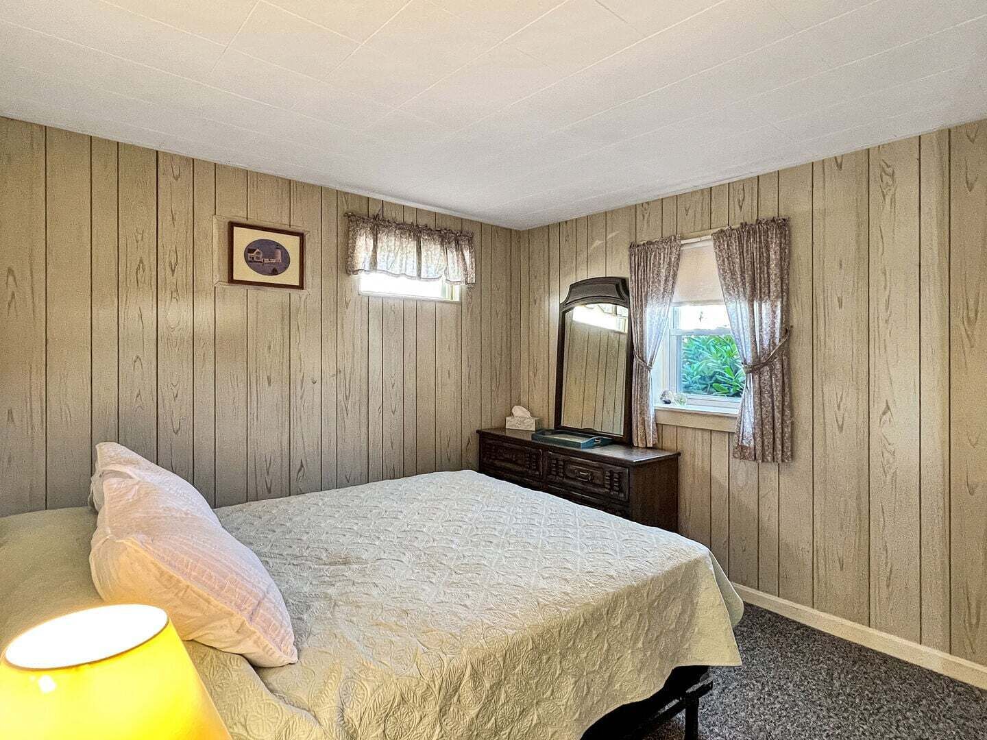 Quarto com cama, cômoda, espelho, janela com cortinas e abajur.