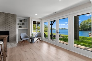 A spacious room with a fireplace, two chairs, a bookshelf, and large windows overlooking a lake. - Fox Run LKN (Denver)