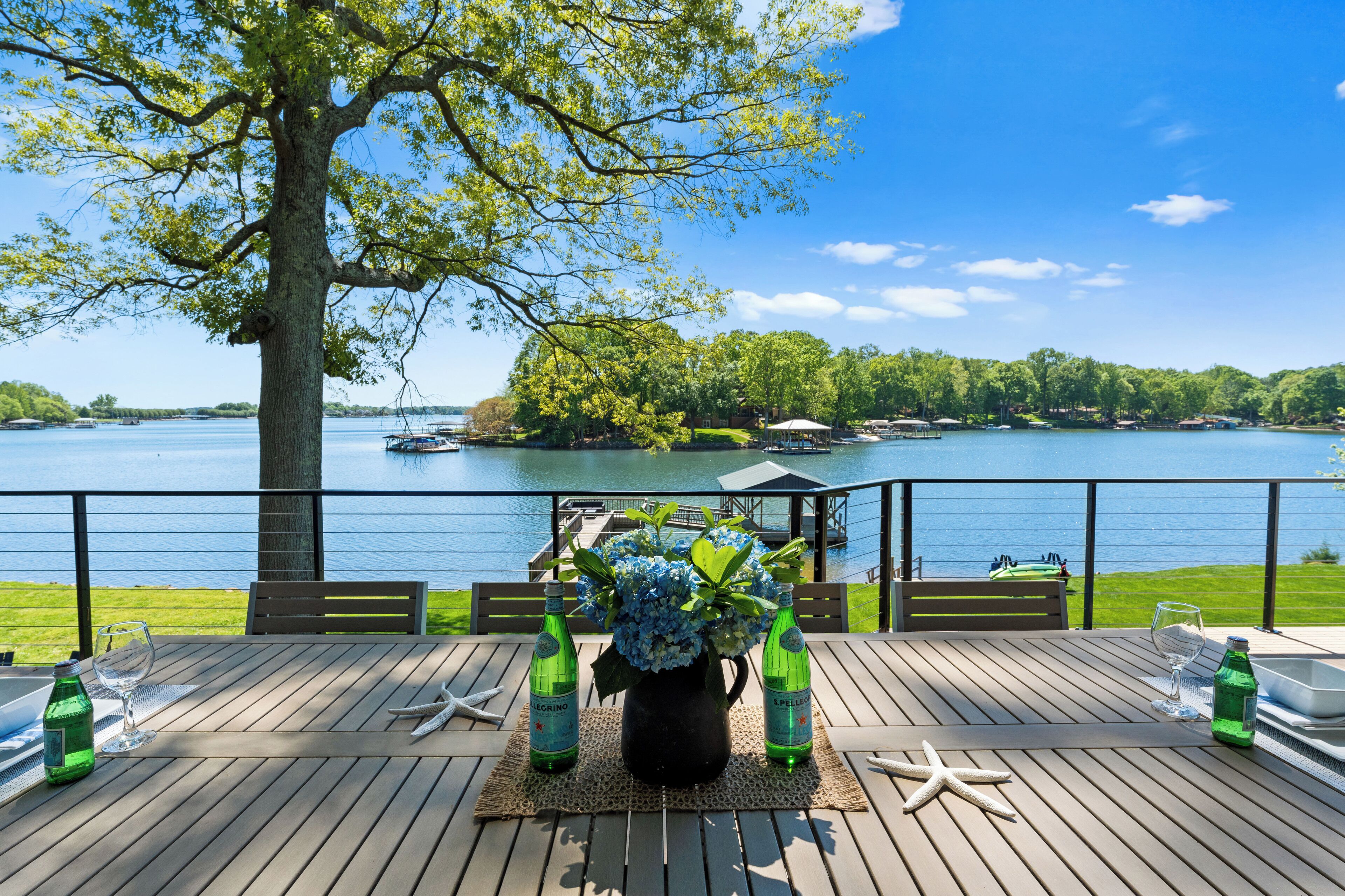 A wooden deck with a table set for two, overlooking a lake with boats and a gazebo.