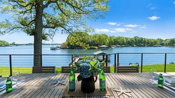 A wooden deck with a table set for two, overlooking a lake with boats and a gazebo.