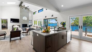 A modern kitchen with a large island, a dining area with a sofa, and a view of a blue body of water through large sliding doors.