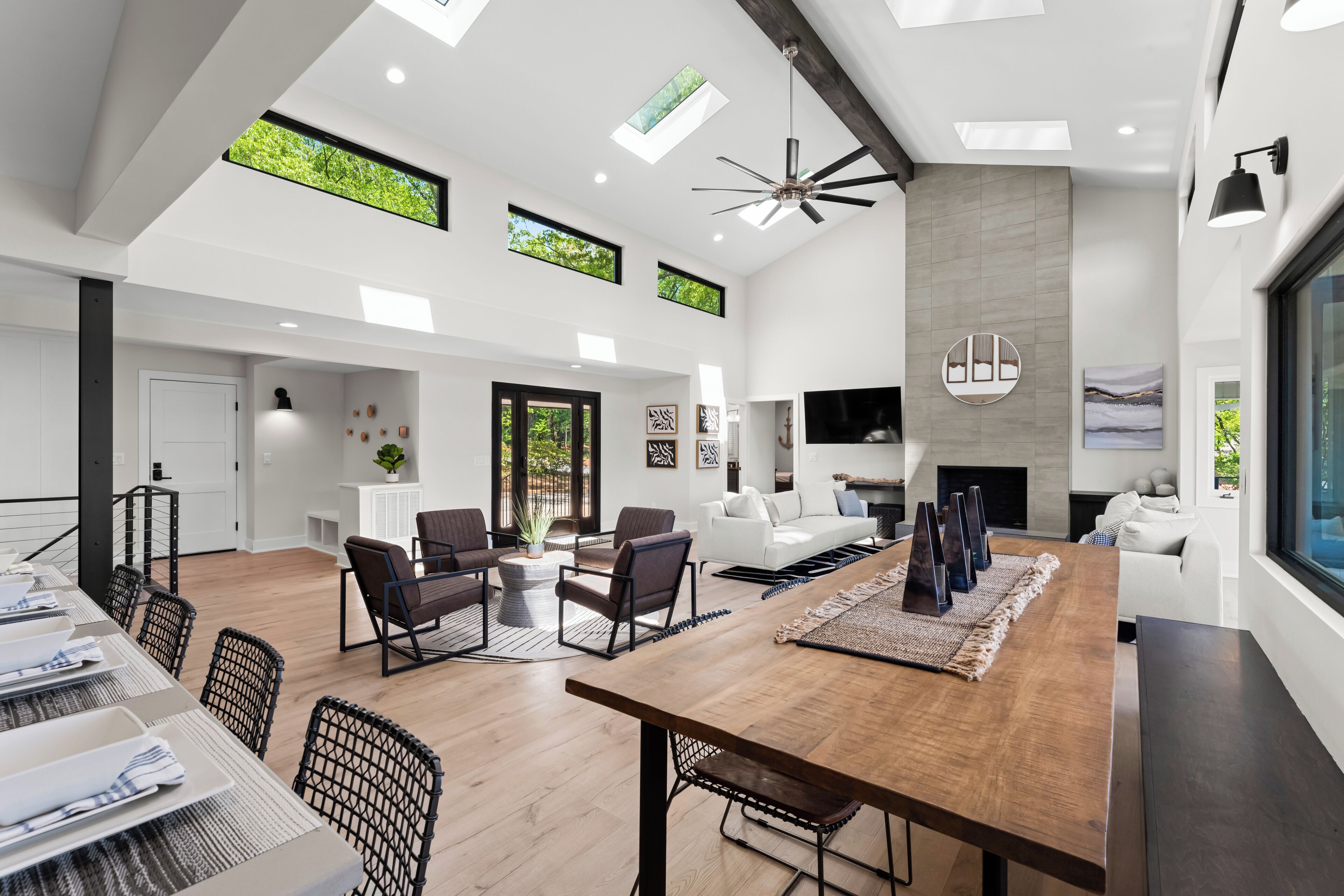 A spacious living area with a large wooden dining table, dark chairs, and a fireplace. There are multiple windows allowing natural light and views of greenery.