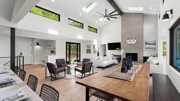 A spacious living area with a large wooden dining table, dark chairs, and a fireplace. There are multiple windows allowing natural light and views of greenery.