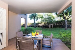 An outdoor patio with a table set for two, surrounded by chairs, and a view of a well-maintained garden and trees. - Beachfront Bliss Walk to Sand, Pool, Restaurants (Kihei)