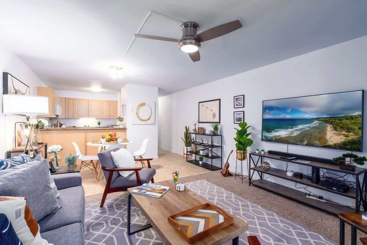 A modern living room with a sofa, coffee table, and TV. There is a kitchen area with wooden cabinets and a dining table.