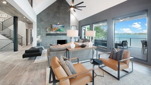 A modern living room with a fireplace, leather sofas, a dining area, and a large window overlooking a body of water. - Sterling on Strawpocket (Mooresville)