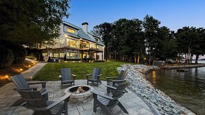 A lakeside patio with Adirondack chairs and a fire pit, overlooking a house with a large deck and a view of the water. - Sterling on Strawpocket (Mooresville)