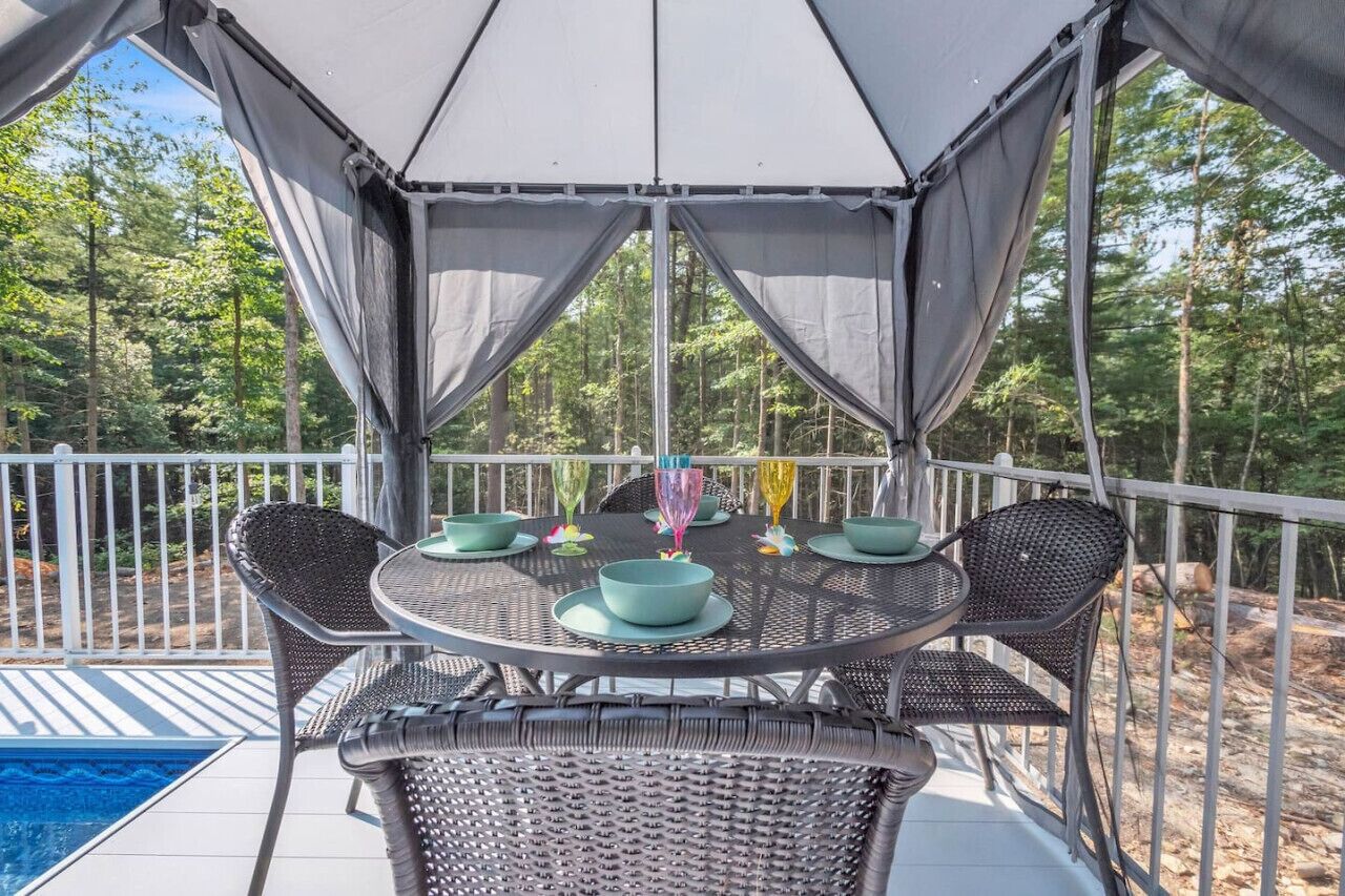 A covered outdoor patio with a round table and chairs, a pool, and a view of a wooded area.