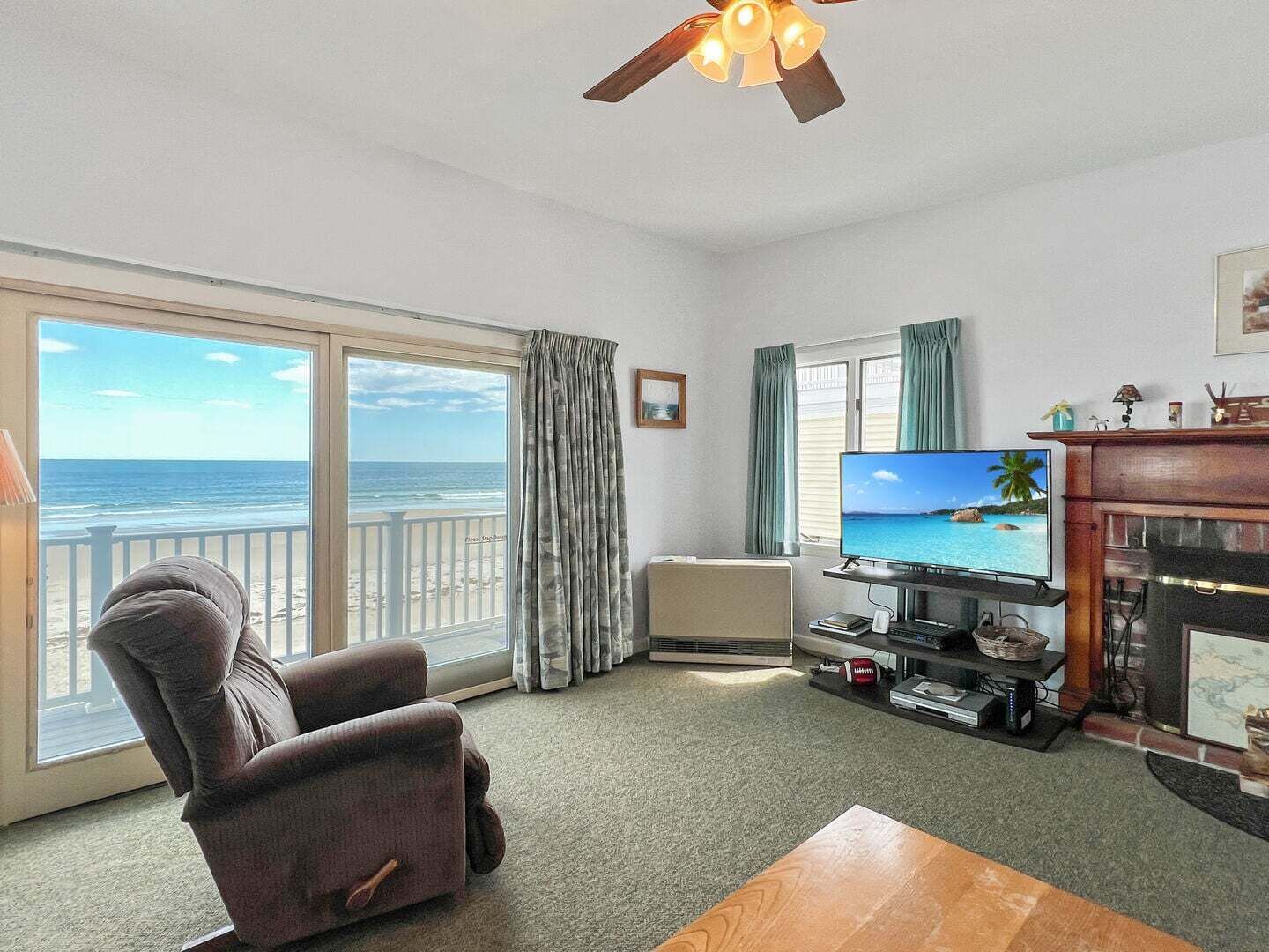 Uma sala de estar com televisão, lareira, janela grande com vista para o oceano, ventilador de teto e poltrona confortável.