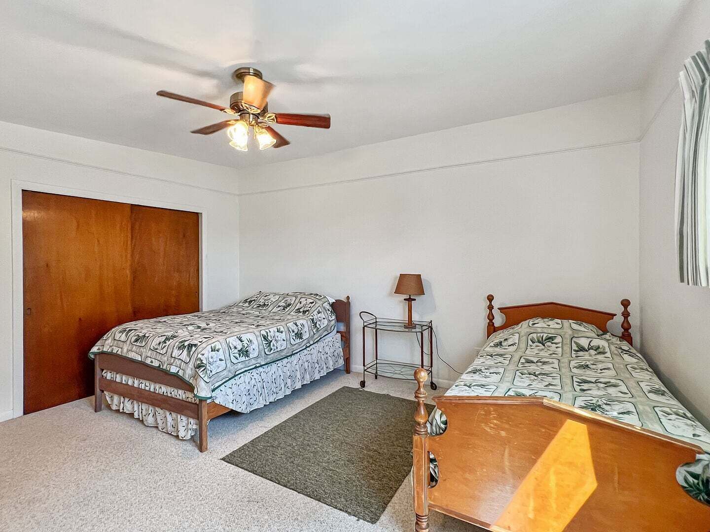 Une chambre avec deux lits simples, un ventilateur de plafond, une table de chevet avec une lampe et une porte.