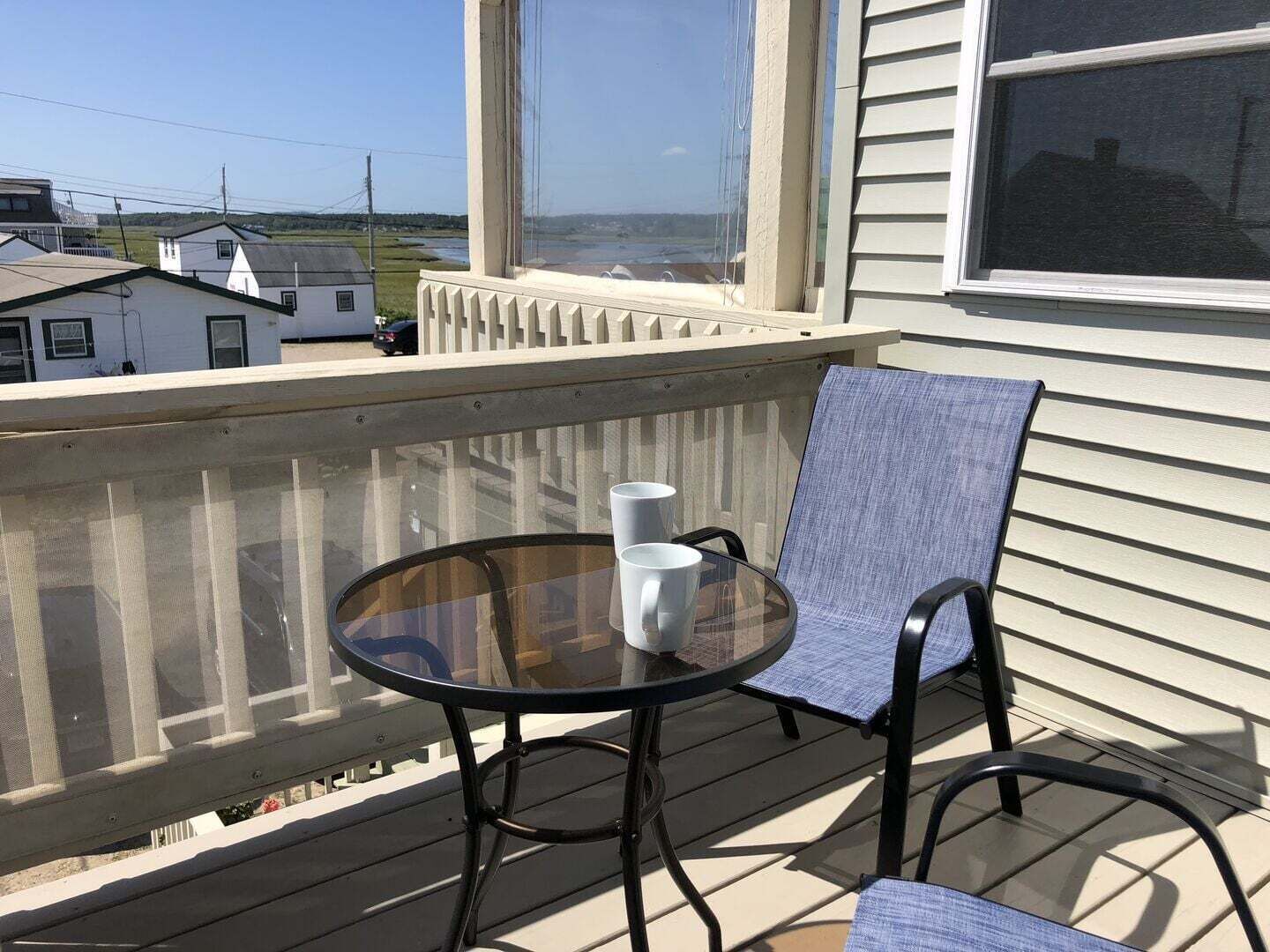 Ein Balkon mit einem Glastisch, zwei Stühlen, einer weißen Tasse und Blick auf Häuser und eine Straße.