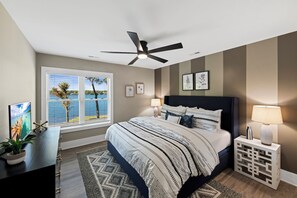 Ein Schlafzimmer mit Bett, Deckenventilator, Fenster mit Jalousien, Nachttisch mit Lampe und Wanddeko.