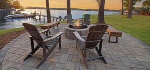 Área de estar externa com cadeiras Adirondack e lareira, com vista para um lago com barcos e cais.
