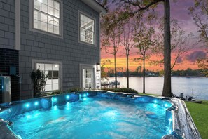 Uma banheira de hidromassagem com vista para um lago e para o pôr do sol, uma casa com janelas amplas e um píer ao fundo.