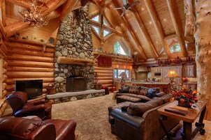 A log cabin interior with a stone fireplace, leather furniture, and a kitchen area.
