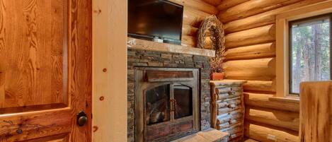 A log cabin interior with a stone fireplace, a television mounted above it, and a window with a view of trees.