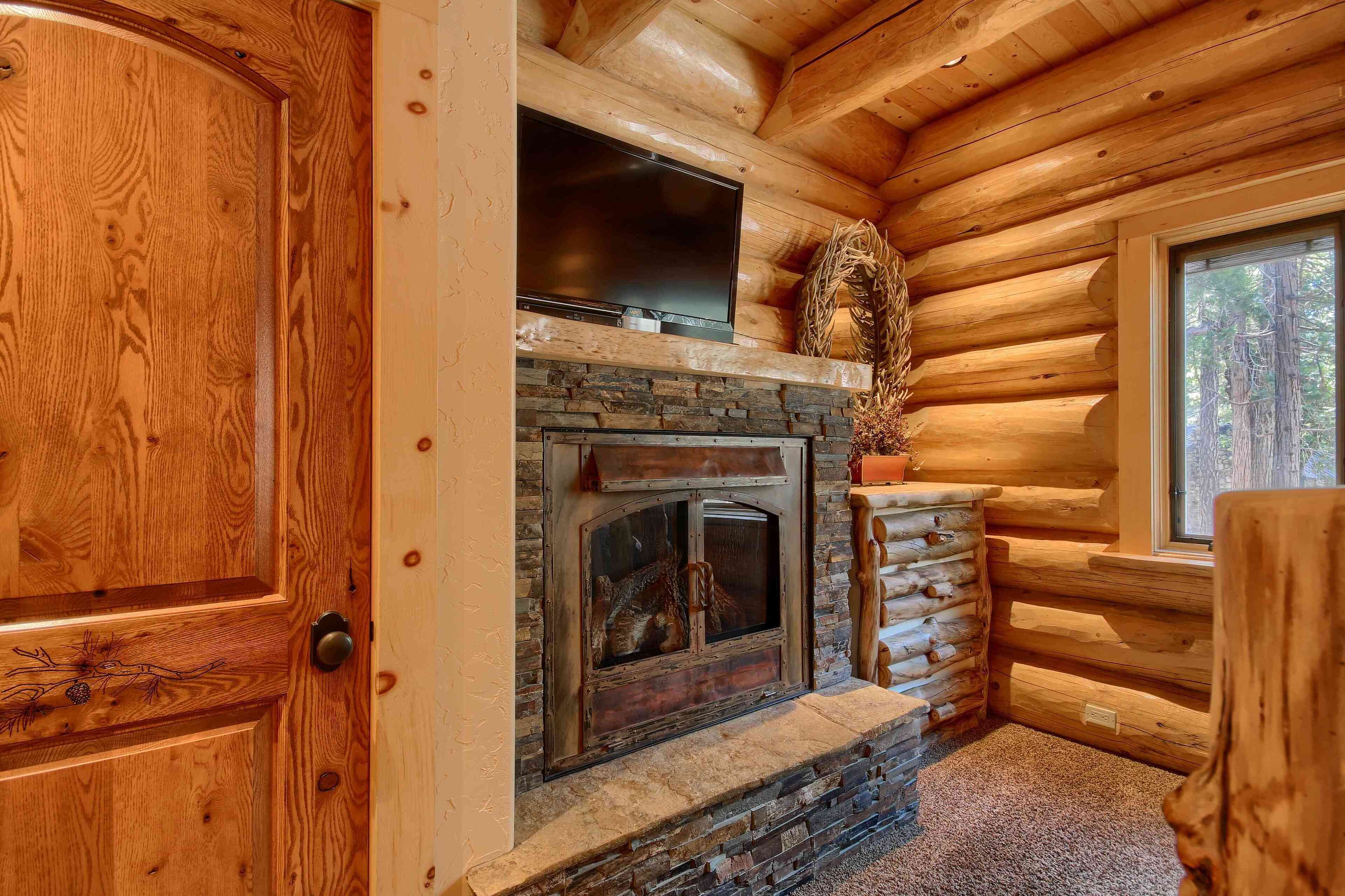 Interior de una cabaña de troncos con chimenea de piedra, un televisor de pantalla plana montado encima y una ventana con vista a árboles.