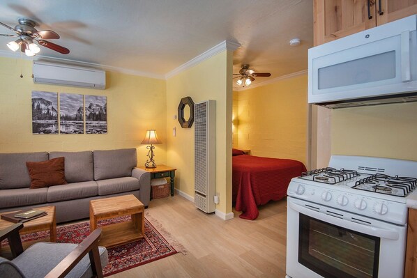 A compact living space with a sofa, a small table, a red bed, a kitchenette with a stove, and a ceiling fan. - Yosemite's Riverfront Retreat (Yosemite National Park)