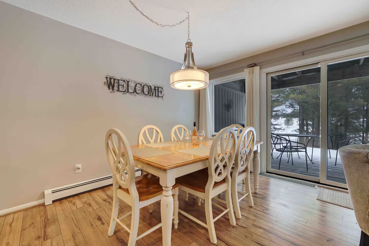 A dining area with a wooden table and white chairs, a 'WELCOME' sign on the wall, and a view of trees through large windows.