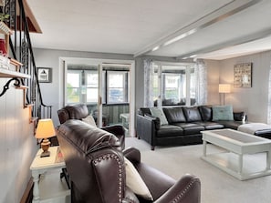 Un salon avec un canapé en cuir, une table basse blanche et une lampe. On y trouve un escalier orné d’une enseigne Coca-Cola, ainsi qu’une grande fenêtre avec des stores.