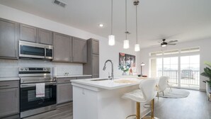 A modern kitchen with stainless steel appliances, a white island with bar stools, and a large window providing natural light. - Luxurious One Bedroom Whitestown (Whitestown)