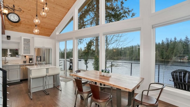 A modern kitchen with large windows overlooking a lake, wooden flooring, and a dining area with metal chairs. - Lakefront Escape Fire Pit, Pets & Amazing Views (Stanwood)