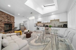 A modern living room with a brick fireplace, a glass dining table, and a sofa. - Marbella Lane - Your Cozy Haven 2BR Retreat (West Covina)