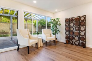 A spacious room with two white armchairs, a small round table, and a large window with a view of greenery. - Marbella Lane- Cozy 4BR Oasis Near Nature Trails (West Covina)