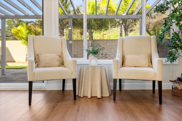 A room with two beige armchairs, a small round table, and a view of greenery through large windows. - Marbella Lane- Cozy 4BR Oasis Near Nature Trails (West Covina)