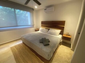 A modern bedroom with a large bed, wooden headboard, bedside tables, and a window with a view of trees. - Beautiful 2 Bedrooms in Parques Zama Torre 2 (Tulum)