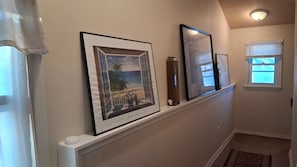 Um corredor com um quadro de uma paisagem de praia, um vaso alto e uma janela com persianas.