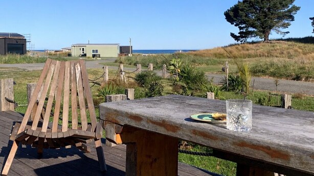 Une terrasse en bois avec une table de pique-nique et une chaise en bois, donnant sur une zone herbeuse avec un arbre et un bâtiment visible au loin.