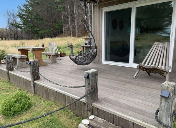 A wooden deck with outdoor furniture, including a hanging chair and a bench, leading to a glass door entrance. - The Beach Apartment (Flat Point)