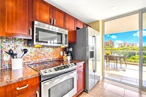 A kitchen with wooden cabinets, a stainless steel oven, a microwave, and a refrigerator. There is a tiled backsplash and a window leading to a balcony with a view of buildings and greenery. - Honua Kai | Ocean View 2 BR | Car Incl. w/ 6+ Nights | HKK-434 by KBM (Lahaina)