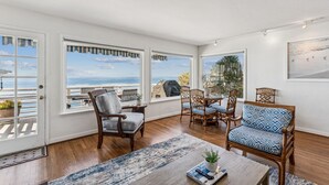 Amplia habitación con grandes ventanales que ofrecen vistas al océano, piso de madera y una combinación de muebles de madera y tapizados.