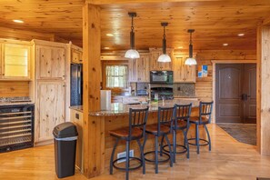 A wooden cabin interior with a kitchen area, bar stools, and a trash bin. - Bear Bluff Cabin / HOT TUB / EV Charge / Sleeps 10 / 3k sq ft / 10 acres (De Soto)