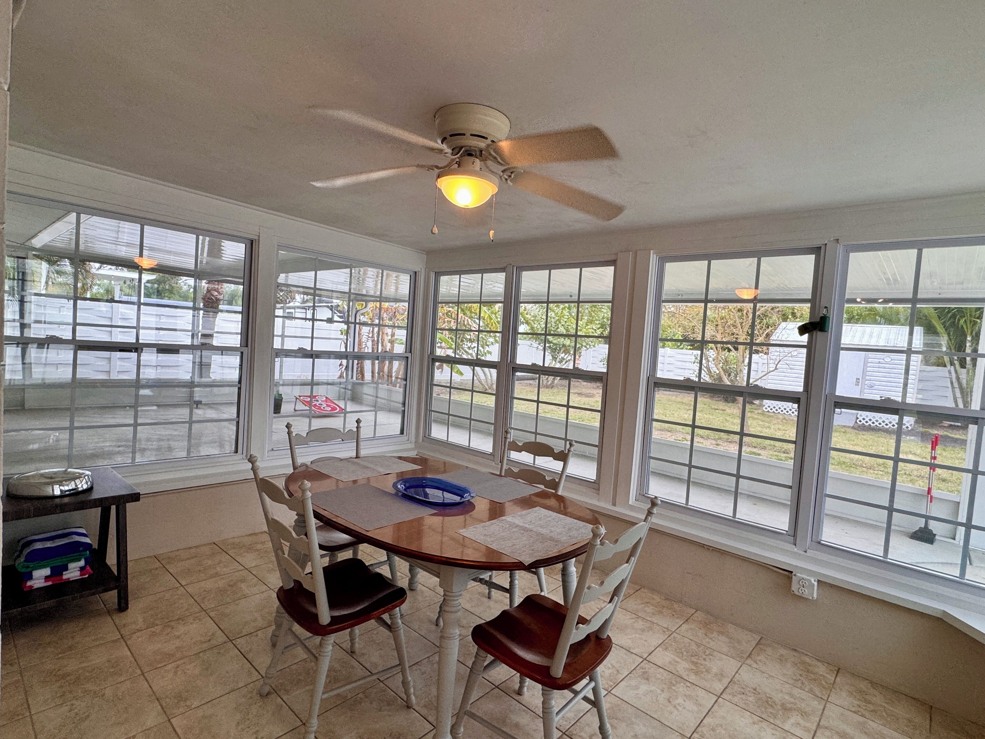 A sunroom with a ceiling fan, large windows, a dining table with chairs, and a small shelf with folded clothes.