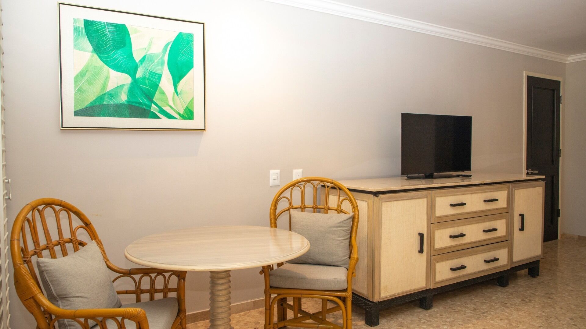 A room with a round table, two wicker chairs, a television on a cabinet, and a framed picture on the wall.