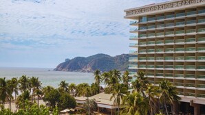 A hotel with multiple floors, balconies with glass railings, and a view of the ocean and a mountain in the background. - Princess Mundo Imperial All Inclusive (Acapulco)