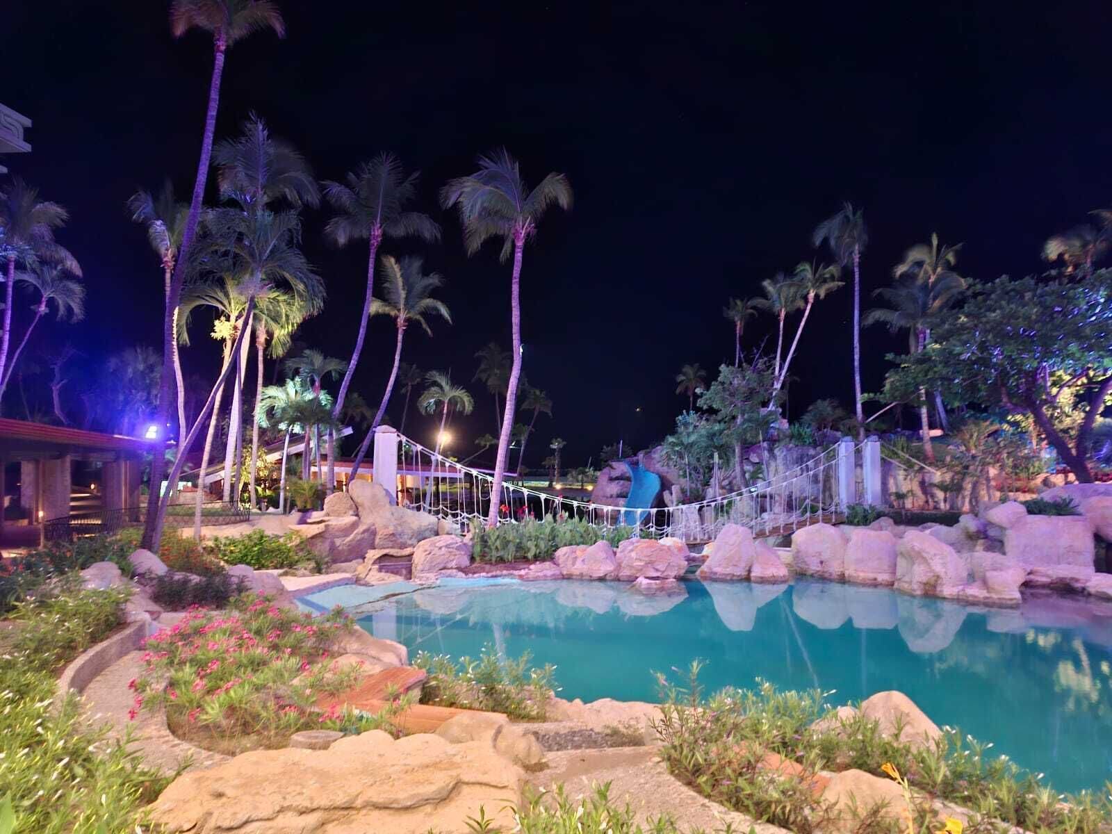 Zona de piscina d'un hotel de nit, amb palmeres, un pont de corda i una cascada.