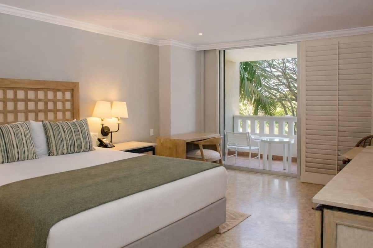 A hotel room with a large bed, a desk, and a balcony with a view of palm trees.