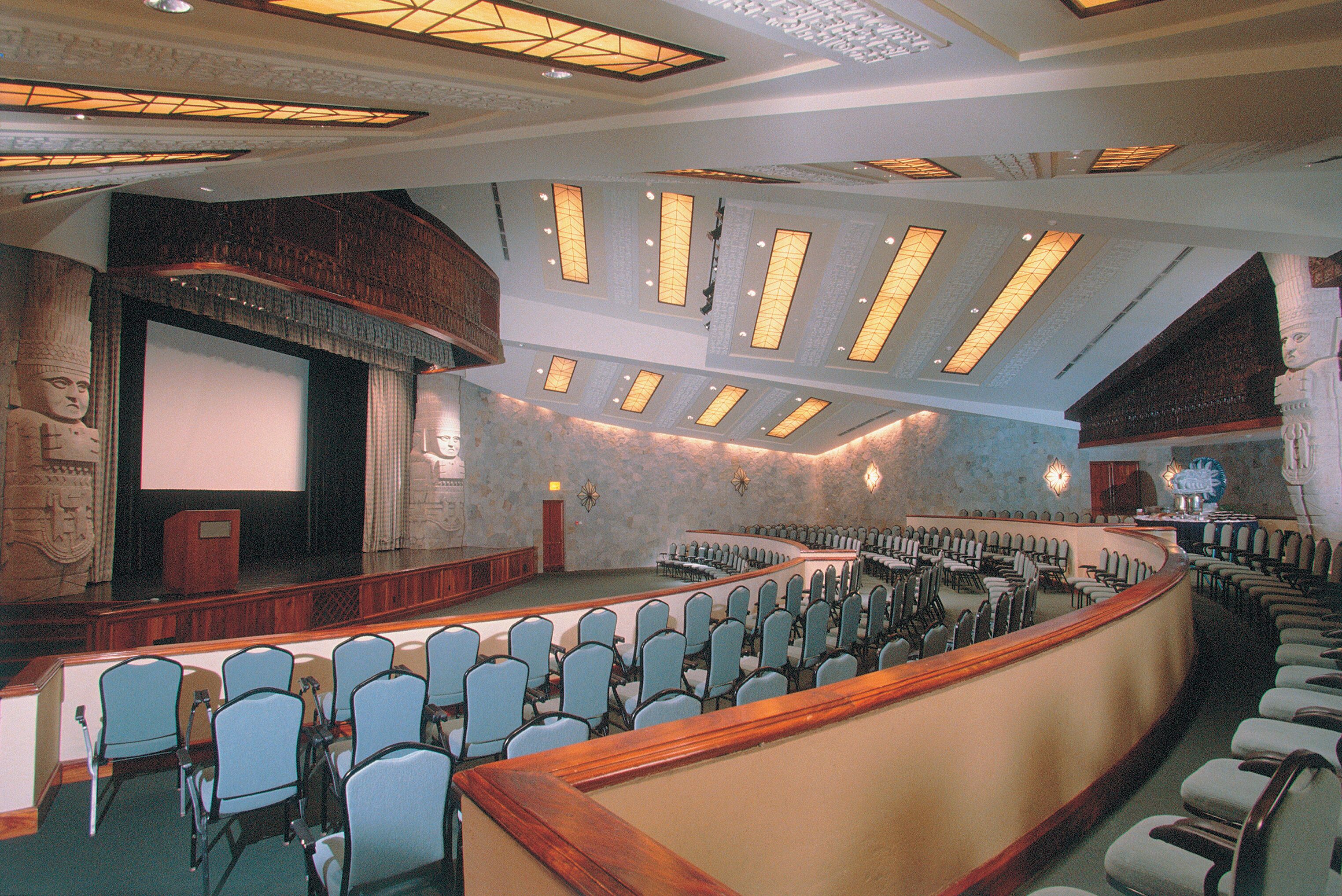 Um amplo auditório com assentos em níveis, palco e uma grande tela.