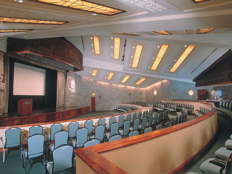 Um amplo auditório com assentos em níveis, palco e uma grande tela.