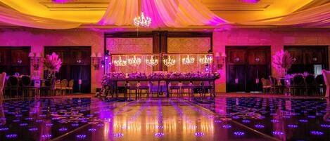 Un salón de banquetes con una larga mesa dispuesta para un evento formal, rodeada de sillas y decorada con arreglos florales.