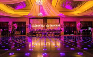 Un salón de banquetes con una larga mesa dispuesta para un evento formal, rodeada de sillas y decorada con arreglos florales.