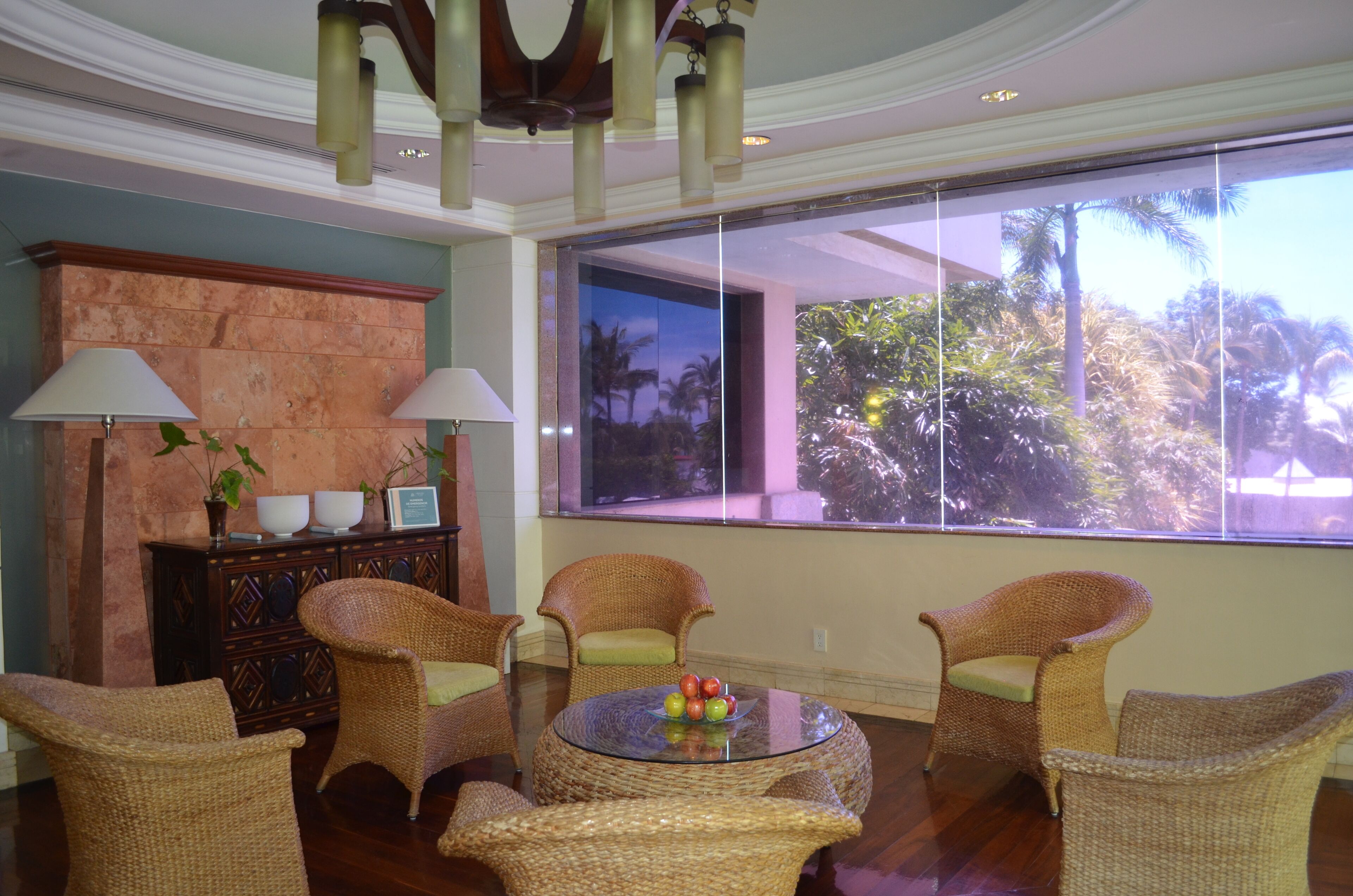 A room with wicker furniture, a glass coffee table with fruit, a fireplace, and a large window with a view of palm trees.